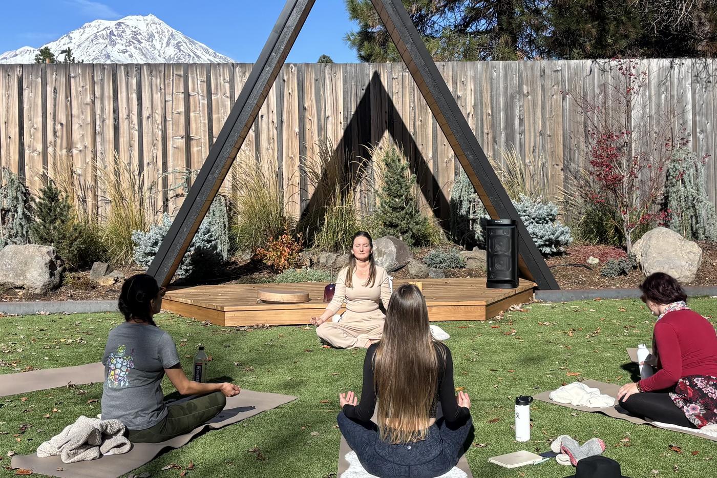 Anya seated beneath a wooden A-frame, leading women in meditation, with snow-capped Mount Shasta behind