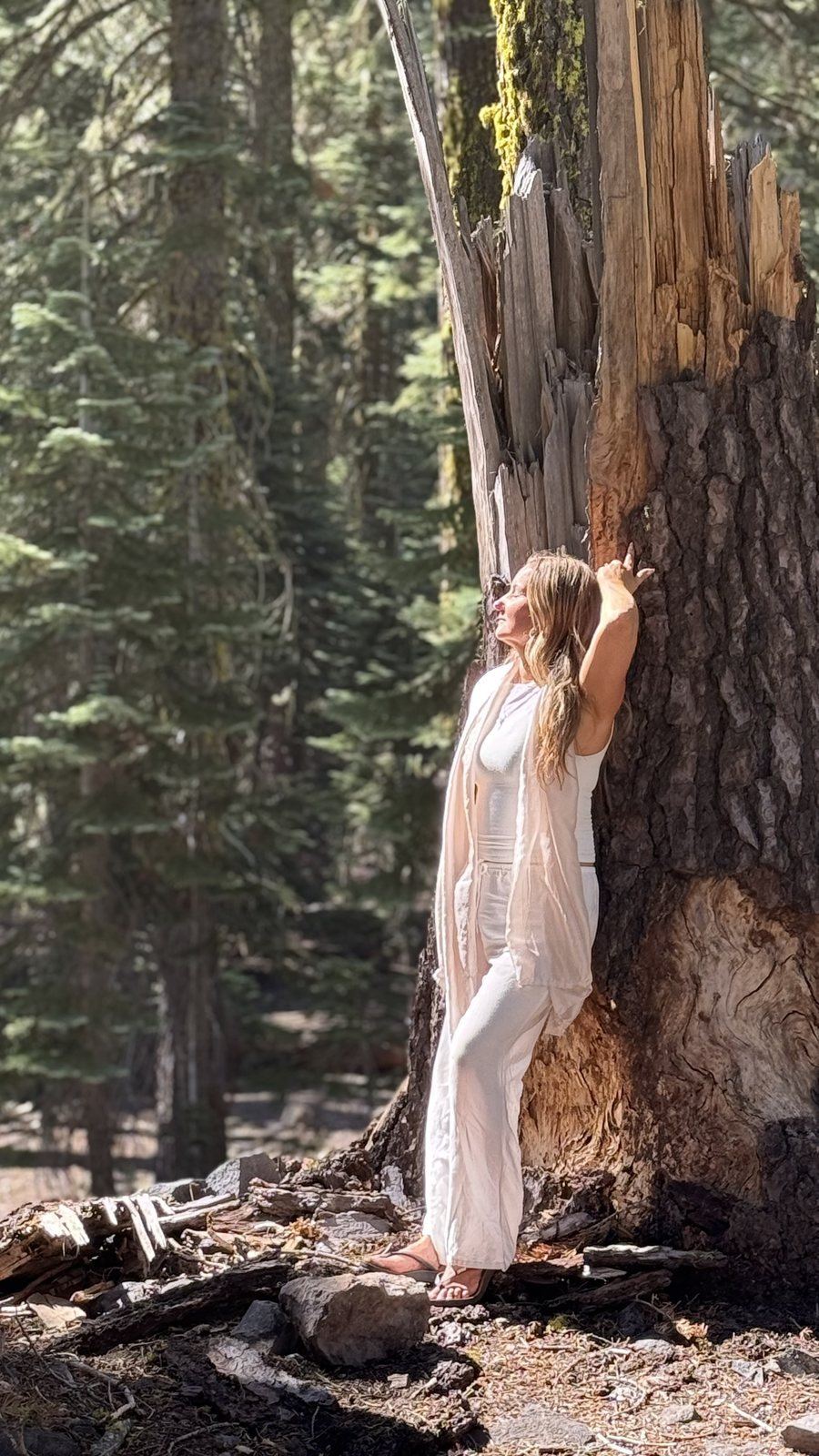 Anya in white standing at the base of an old tree on the mountain